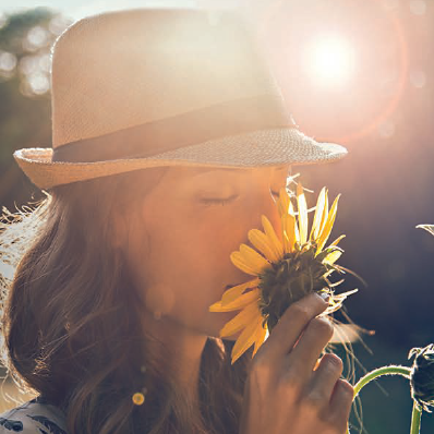 Eine Frau mit Strohhut riecht mit geschlossenen Augen an einer Sonnenblume.